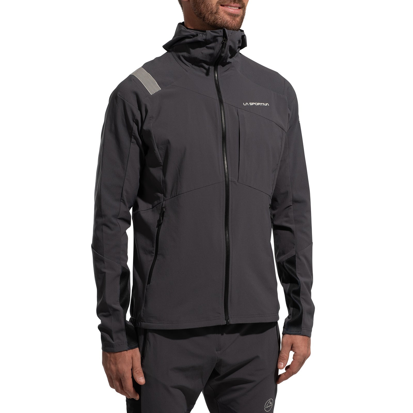 A man wears the La Sportiva Aequilibrium Speed Jacket Men's (2025) in dark gray with matching pants, made from breathable fabric. The jacket has a zipper and reflective shoulder details; he stands relaxed against a white background.