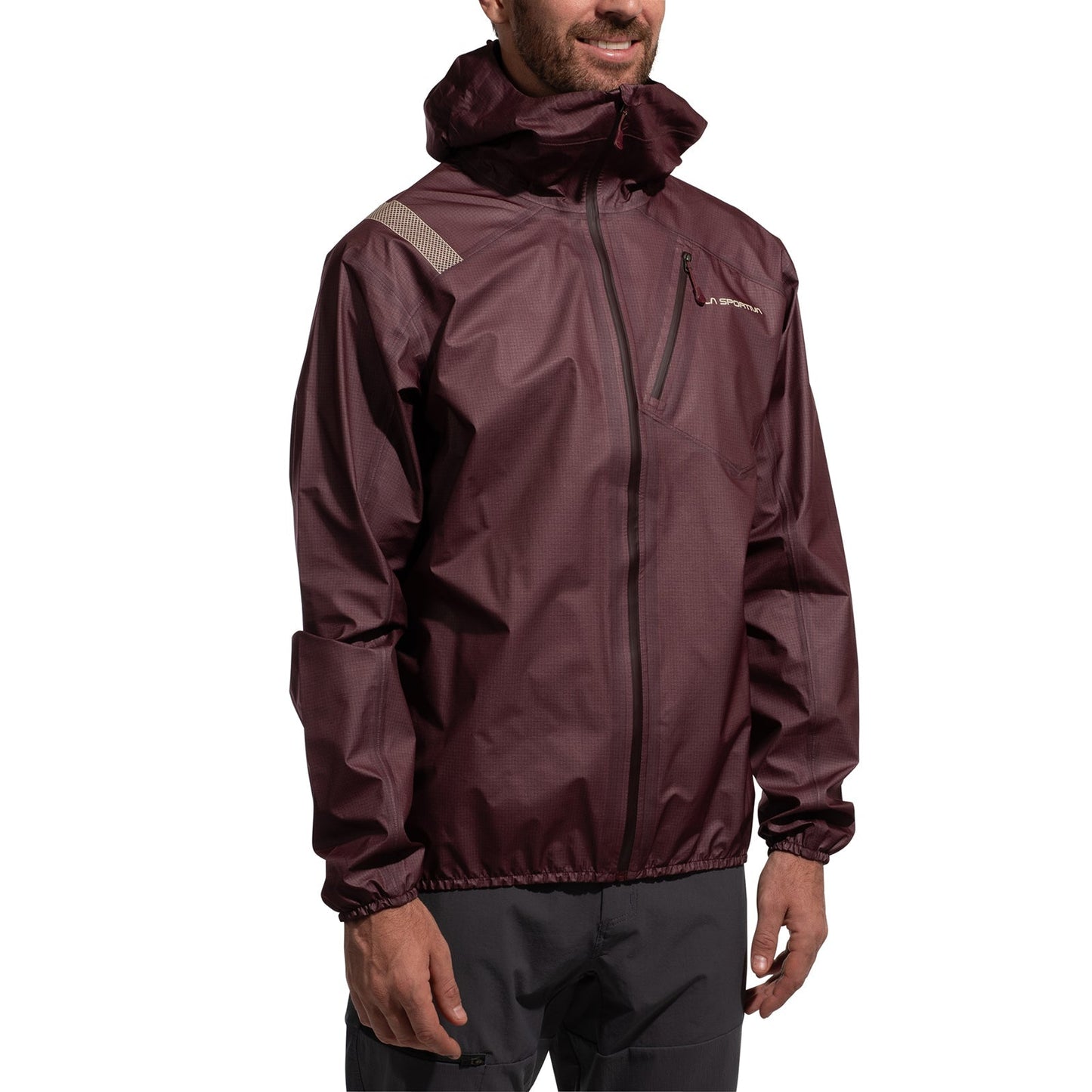 A person is smiling while wearing the La Sportiva Aequilibrium Lite GTX Jacket Men's, a maroon lightweight mountaineering jacket with a front zipper, gray shoulder detail, chest pocket, and elastic cuffs, paired with dark pants.