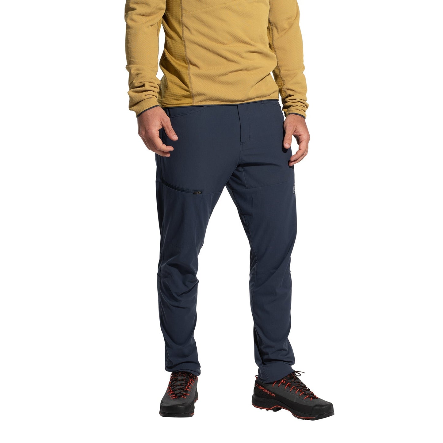 Wearing a mustard long-sleeve shirt, La Sportiva Machina Tech Pants Men's (2025) in navy blue, and black hiking shoes with red laces, a person stands against a white background. The image shows them from shoulders to feet.