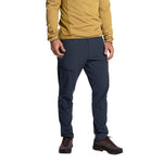 Wearing a mustard long-sleeve shirt, La Sportiva Machina Tech Pants Men's (2025) in navy blue, and black hiking shoes with red laces, a person stands against a white background. The image shows them from shoulders to feet.