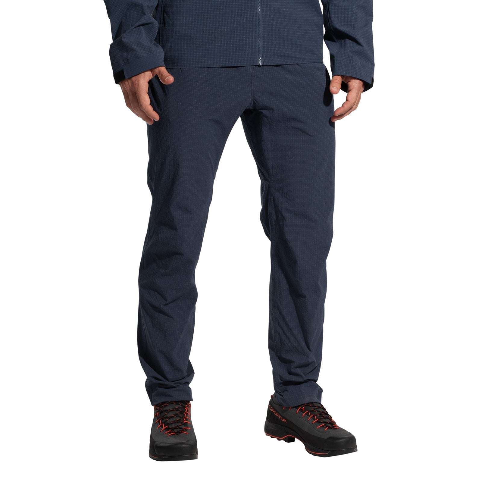 A person wears La Sportiva Helixir Pants Men's (2025) in dark blue with harness-compatible pockets and black hiking boots with red laces, standing against a white background. Their face is not visible.