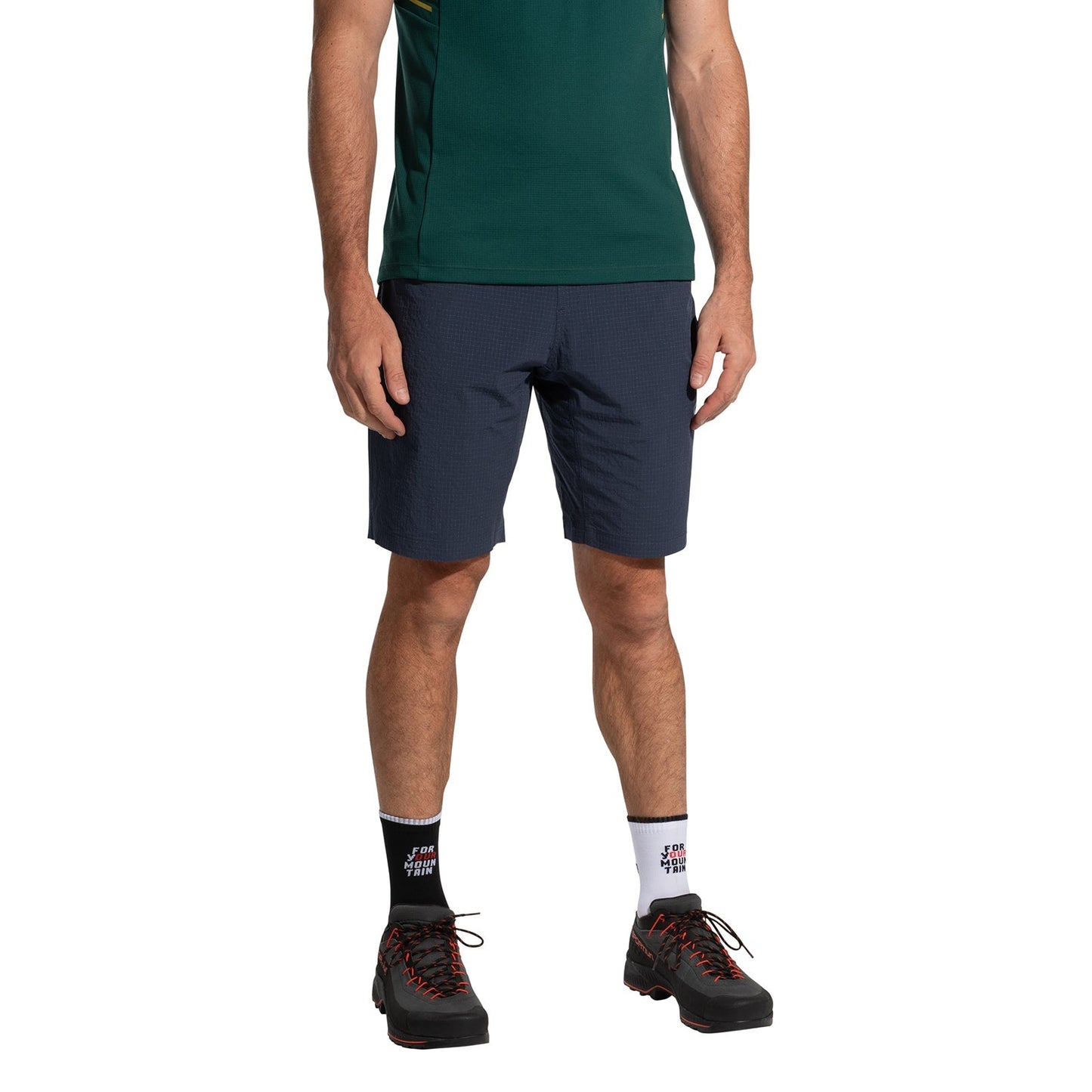 A man in a dark green shirt, La Sportiva Helixir Shorts Men's (2025), black hiking shoes with red laces, and mismatched socks stands against a white background—ready for adventure. His face is not visible.