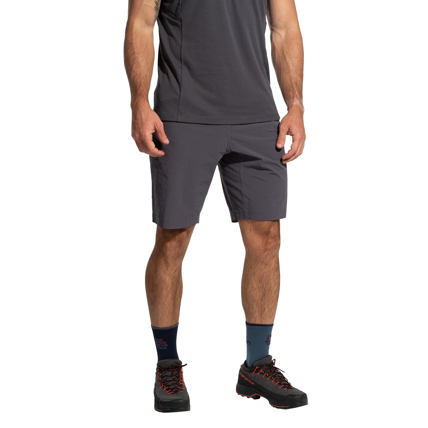 A man stands against a white background, wearing La Sportiva Helixir Shorts Men's (2025) in dark gray, a matching athletic shirt, navy socks, and black hiking shoes with red accents; his head is not visible, channeling a big wall adventure vibe.
