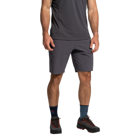 A man stands against a white background, wearing La Sportiva Helixir Shorts Men's (2025) in dark gray, a matching athletic shirt, navy socks, and black hiking shoes with red accents; his head is not visible, channeling a big wall adventure vibe.