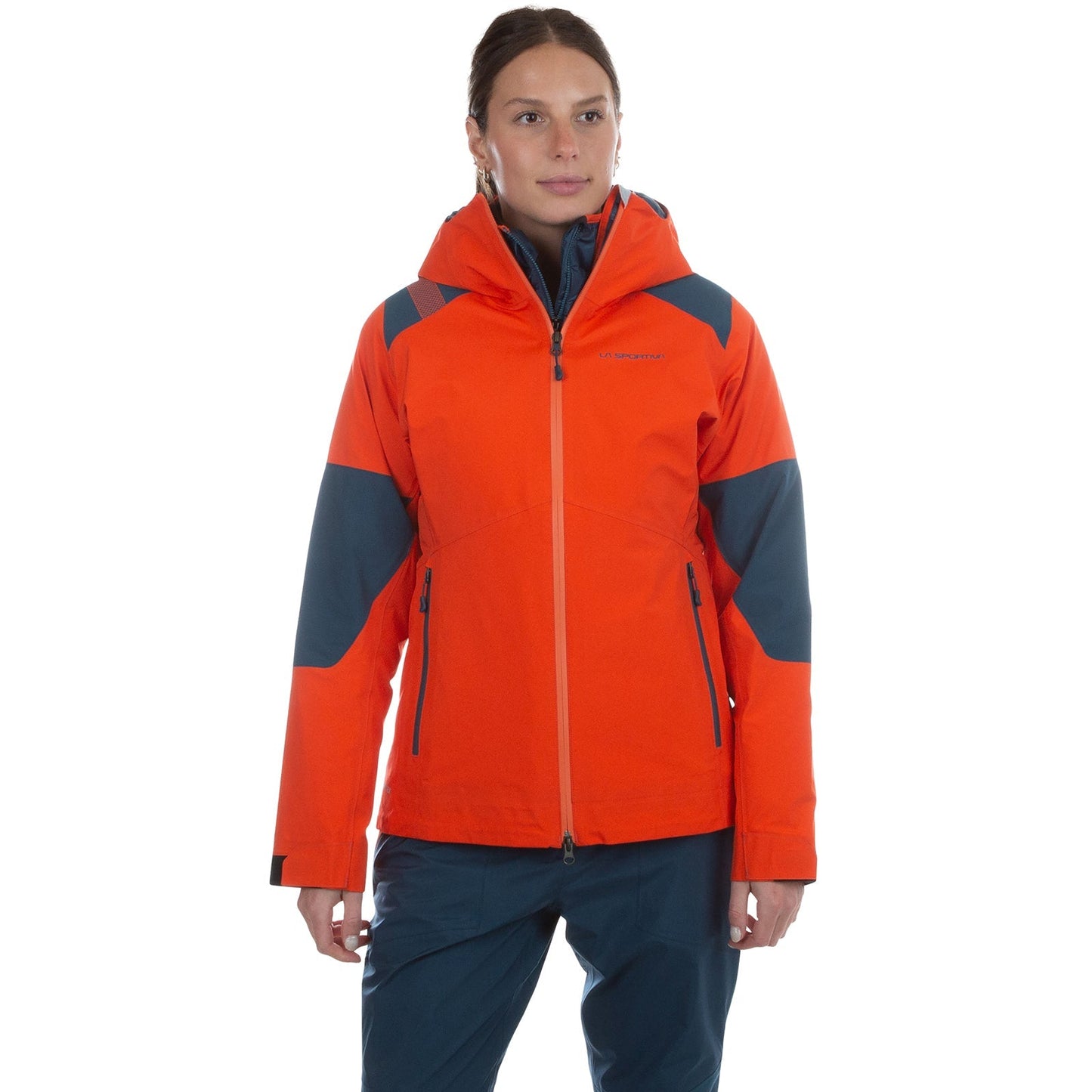 A person stands facing forward against a white background, wearing the La Sportiva Roseg GTX Jacket Women's in orange and gray, paired with blue pants.