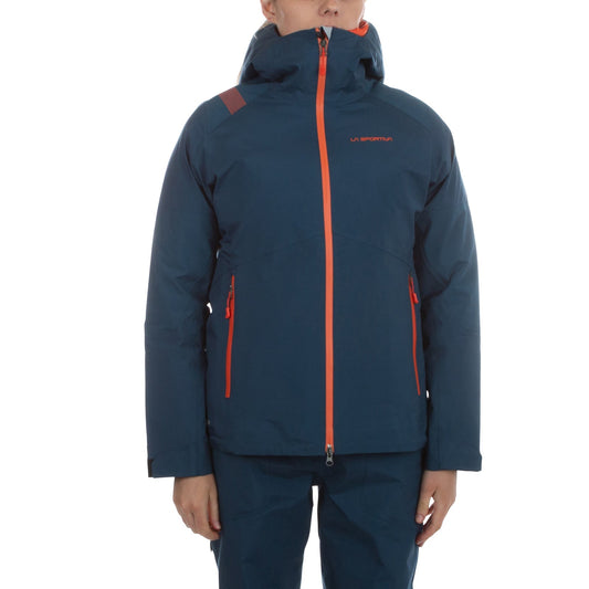 A woman wearing La Sportiva's Roseg GTX Jacket in dark blue with orange zippers and matching pants stands against a plain white background. The high-collar GORE-TEX jacket features an upraised hood for enhanced weather protection.
