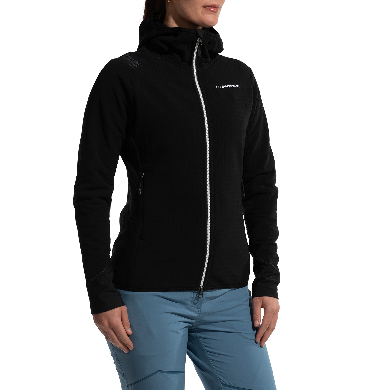 A woman wears the La Sportiva Bristen Thermal Hoody—a black fleece jacket with a hood, white front zipper, and chest logo—paired with light blue pants. She stands relaxed, facing slightly left. This jacket is ideal for outdoor activities.