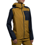 A woman wears the La Sportiva Wall Breeze Stretch Jacket in yellow and navy, featuring a helmet-compatible hood, front zipper, and chest pocket, standing against a white background.