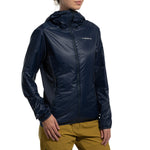 A person wearing the La Sportiva Aequilibrium Lite Insulation Jacket Women's (2025), a dark blue, breathable, warm jacket with a front zipper, and light brown pants stands against a white background. Only the upper body and part of the face are visible.