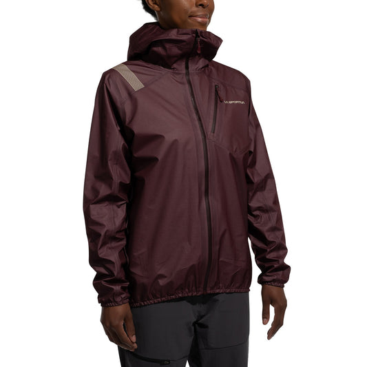 A woman is wearing the La Sportiva Aequilibrium Lite GTX Jacket, a maroon lightweight mountaineering jacket featuring a chest pocket and light gray shoulder detailing, standing against a plain white background.