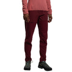 A person stands facing forward, visible from shoulders down against a white background, wearing La Sportiva Machina Tech Pants Women's (2025) in maroon, paired with a long-sleeve pink top and black hiking shoes.