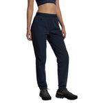 A woman wears La Sportiva Helixir Pants Women's (2025) in navy blue—made with durable Spectra® fibers—and a matching sports bra, standing hands in pockets. She completes the look with black hiking shoes featuring colorful laces.