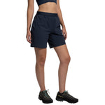A woman in a navy blue crop top, La Sportiva Helixir Shorts Women's (2025) made with durable Spectra fibers, and black trail shoes with yellow laces stands against a white background—ready for any big wall adventure.