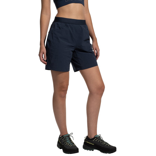A woman in a navy blue crop top, La Sportiva Helixir Shorts Women's (2025) made with durable Spectra fibers, and black trail shoes with yellow laces stands against a white background—ready for any big wall adventure.