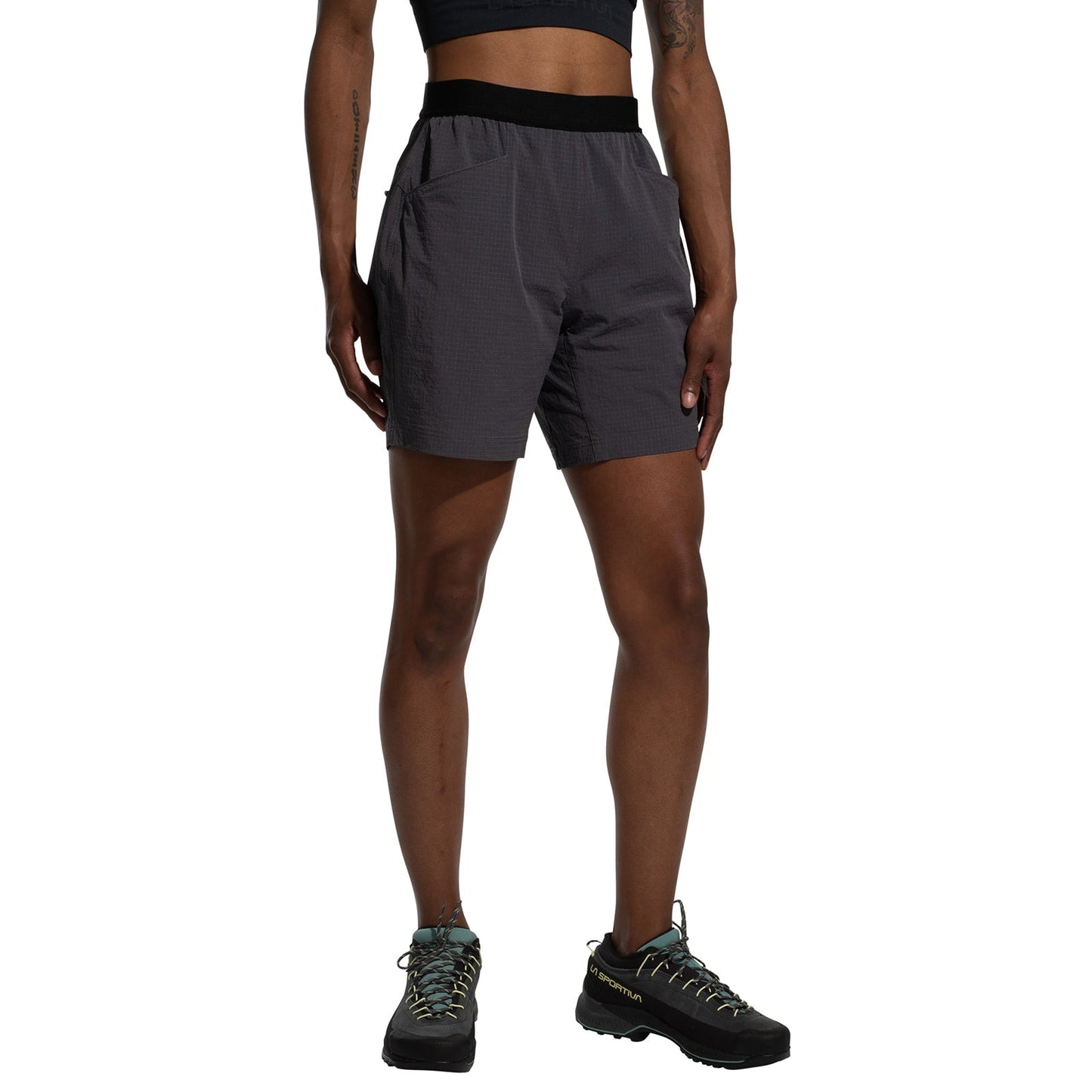 A person wearing a black sports bra, La Sportiva Helixir Shorts Women's (2025) with durable Spectra fibers, and black trail running shoes stands against a white background, hands by their sides, ready for their next big wall adventure.
