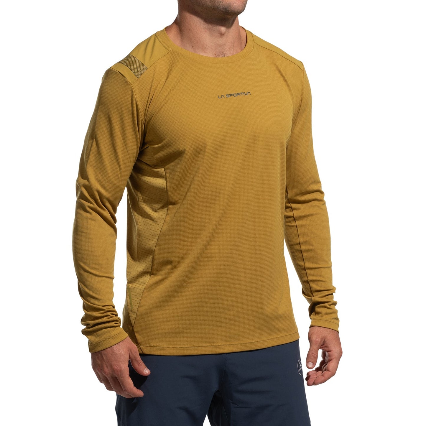 A person wears a mustard yellow La Sportiva Beyond Long Sleeve Men's (2025) top with lightweight Polartec Delta Cooling Fabric and navy pants, standing with arms relaxed against a white background. The head is partially out of frame.