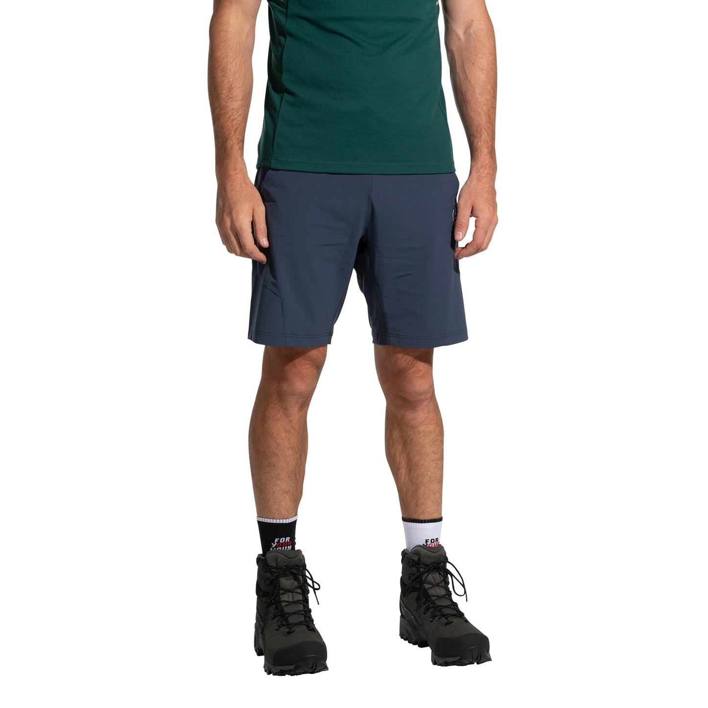 A man wearing a green shirt, La Sportiva Trail Guard Shorts Men's in navy blue, white socks, and black hiking boots stands against a plain white background with his arms relaxed by his sides.