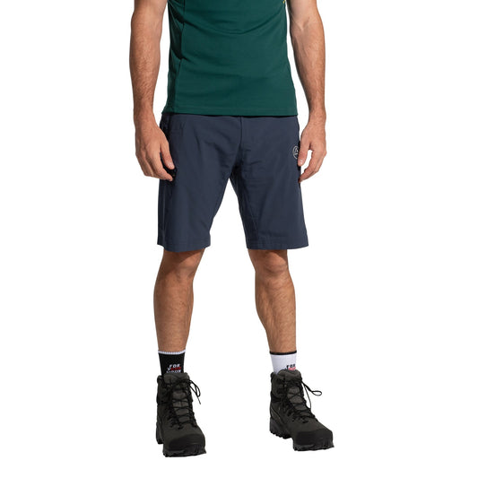 A person stands against a plain white background, wearing a dark green short-sleeve shirt, La Sportiva West Crest Shorts Men's (2025), black hiking boots, and white socks. Their head is cropped out to showcase the outdoor apparel.