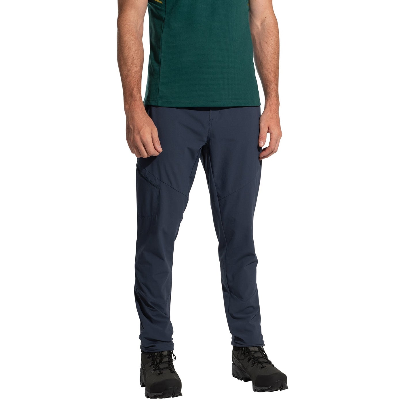 A person wearing a green shirt, La Sportiva West Crest Pants Men's (2025) in dark blue with zippered pockets, and black hiking boots stands against a white background. The person's head is not shown.