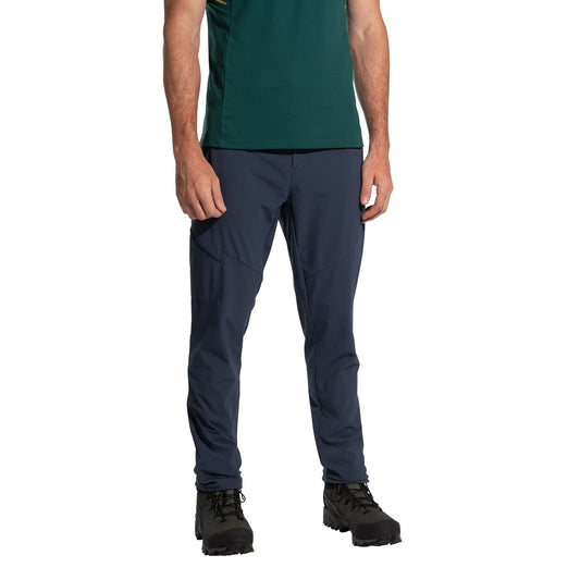 A person wearing a green shirt, La Sportiva West Crest Pants Men's (2025) in dark blue with zippered pockets, and black hiking boots stands against a white background. The person's head is not shown.