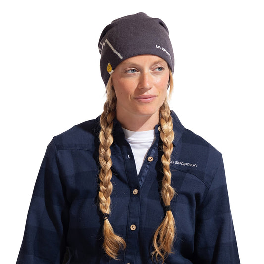 A woman with long blonde braids wears a La Sportiva Woolly Beanie and a navy plaid shirt, both showing the La Sportiva logo. She looks to the side with a neutral expression, ready for cold weather in her comfortable outfit.