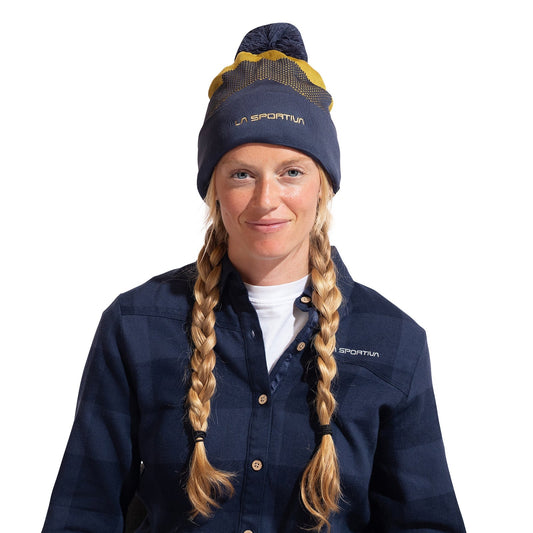 A woman with long blonde braids faces the camera, smiling slightly against a white background. She wears the Knitty Beanie and a blue checkered jacket, both by La Sportiva, showing off her stylish cold-weather look.