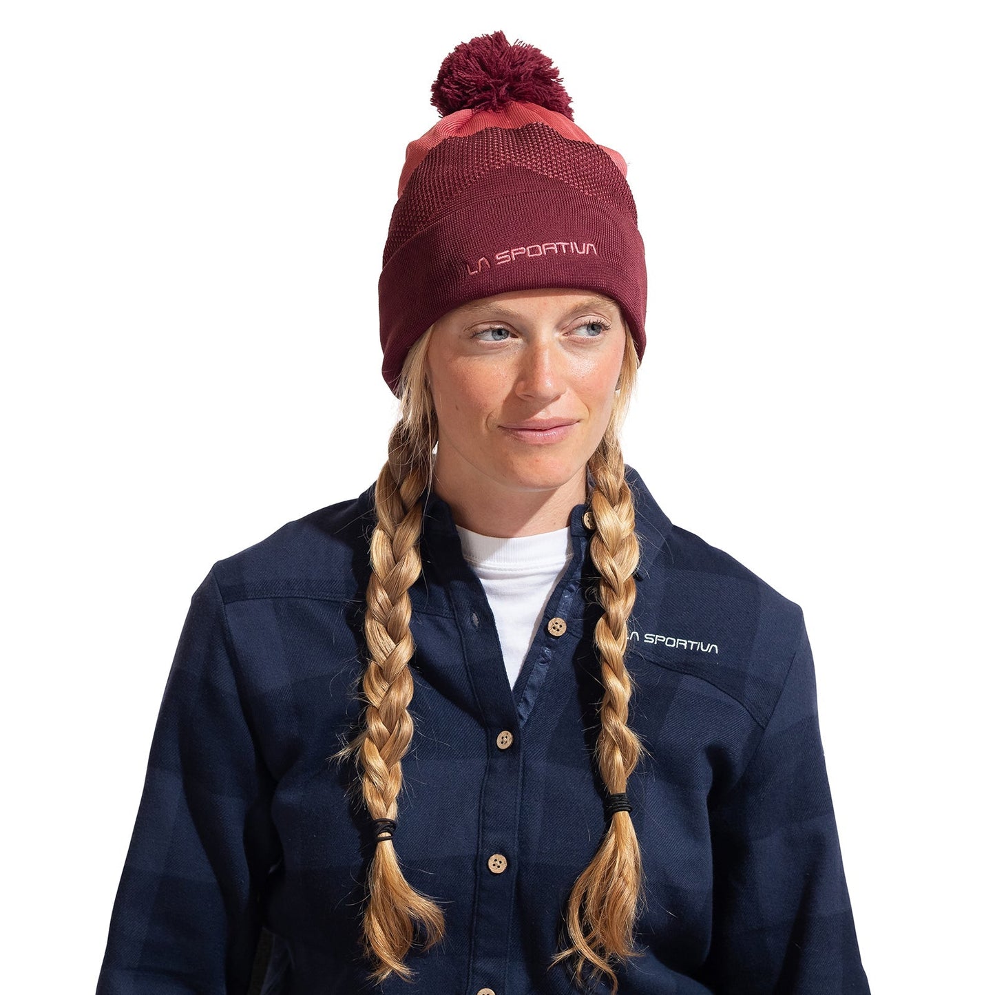 A woman with two blonde braids wears the La Sportiva Knitty Beanie, a maroon and pink knit hat with a pom-pom, and a blue plaid button-up shirt. She looks slightly to the side, smiling softly in this stylish cold-weather look.