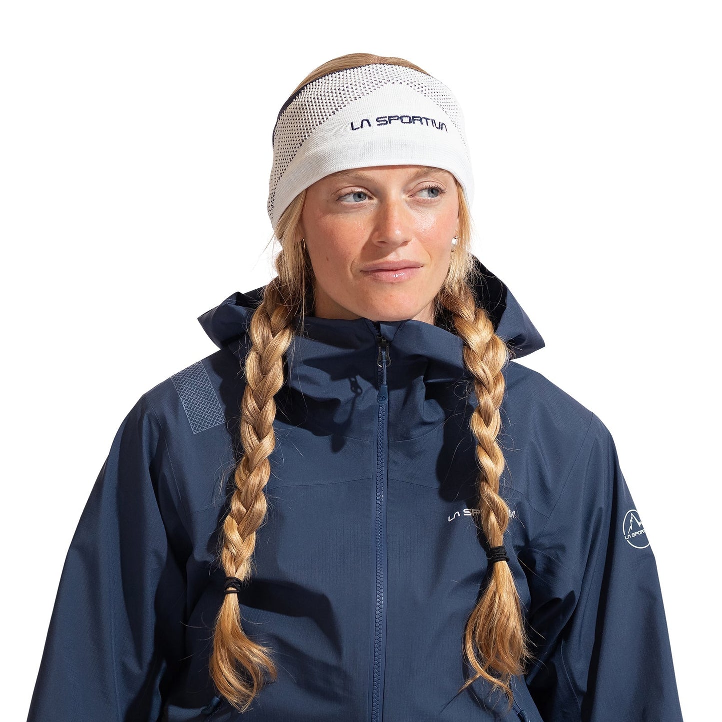 A woman with blonde braids wears a white La Sportiva Knitty Headband ear warmer and a dark blue outdoor jacket, looking slightly to the side against a plain white background.