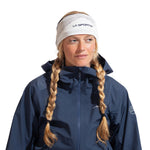 A woman with blonde braids wears a white La Sportiva Knitty Headband ear warmer and a dark blue outdoor jacket, looking slightly to the side against a plain white background.