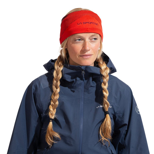 A woman with long blonde braids wears the La Sportiva Knitty Headband as an ear warmer and a dark blue outdoor jacket, perfect for cold weather. She looks slightly to her right against a white background.