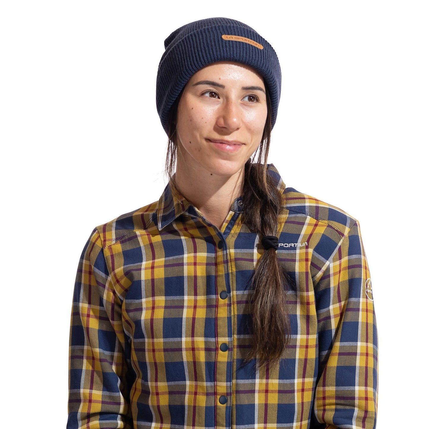 A person with long brown hair in a side braid is smiling and looking to the side, wearing the La Sportiva Boulder Beanie in navy and a yellow, blue, and gray plaid button-up shirt against a white background.