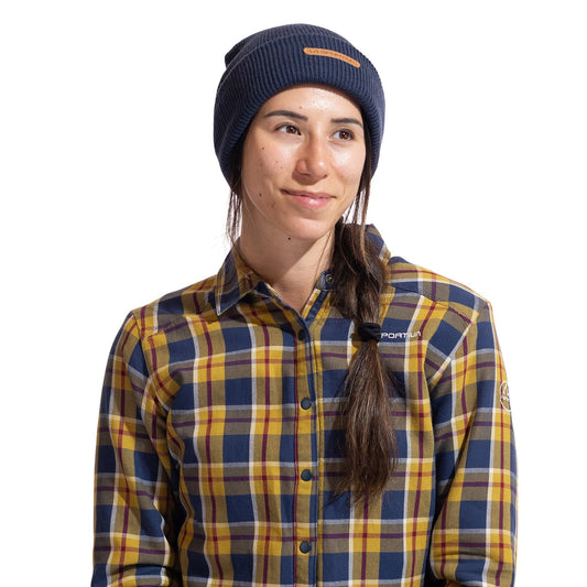 A person with long brown hair in a side braid is smiling and looking to the side, wearing the La Sportiva Boulder Beanie in navy and a yellow, blue, and gray plaid button-up shirt against a white background.