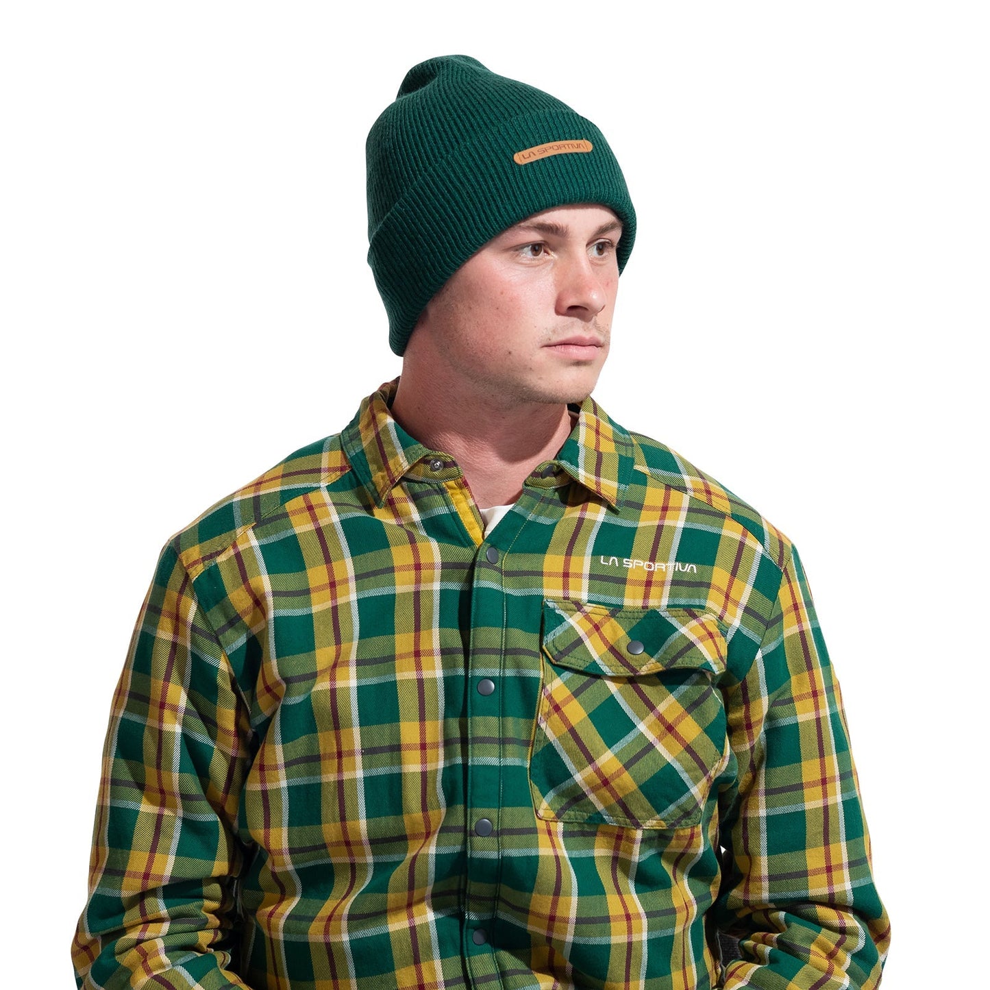 A man in a green La Sportiva Boulder Beanie and a green, yellow, and white plaid button-up shirt with a front pocket looks slightly to the side against a white background.