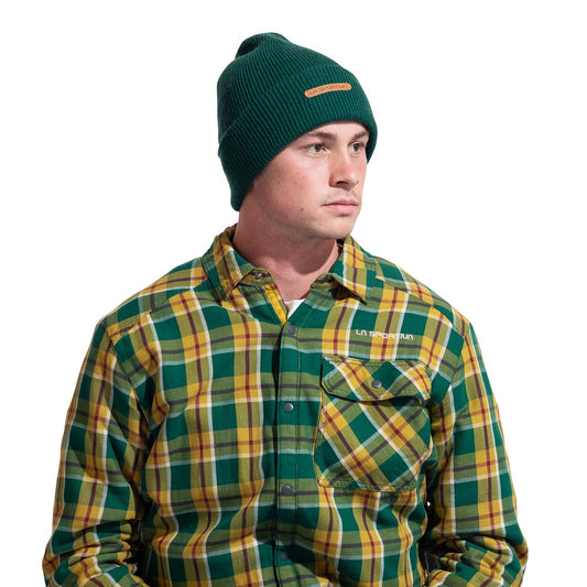 A man in a green La Sportiva Boulder Beanie and a green, yellow, and white plaid button-up shirt with a front pocket looks slightly to the side against a white background.