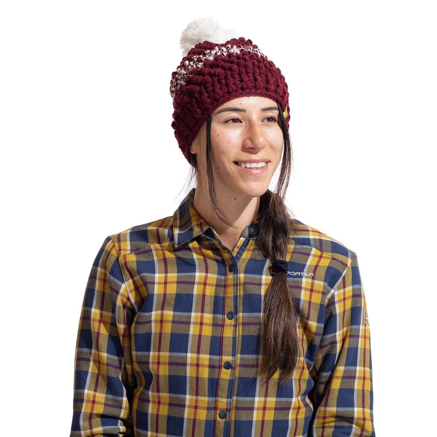 A woman with long dark hair in a side braid wears the La Sportiva Terry Beanie Women's, a maroon winter hat with a white pom-pom, and a yellow-blue plaid shirt, smiling slightly to the side against a white background.