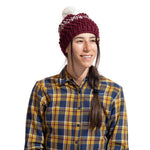 A woman with long dark hair in a side braid wears the La Sportiva Terry Beanie Women's, a maroon winter hat with a white pom-pom, and a yellow-blue plaid shirt, smiling slightly to the side against a white background.