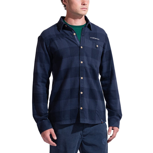 A man wears the La Sportiva Rambler Flannel Shirt Men's, a navy blue plaid cotton-polyester blend with a collar, chest pocket, light buttons, and the LA SPORTIVA logo, standing against a white background.