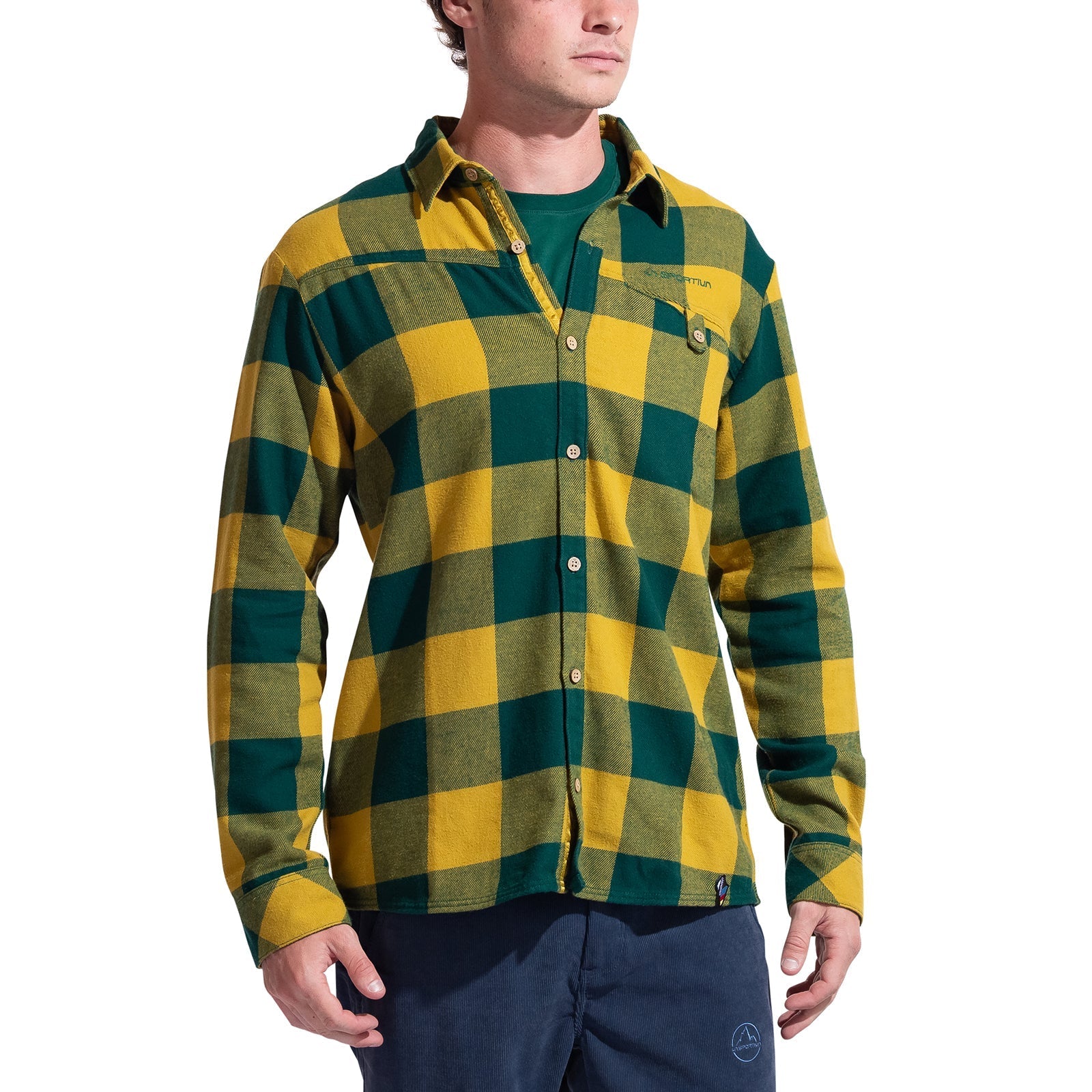 A man wears the La Sportiva Rambler Flannel Shirt in yellow-green plaid with coconut buttons over a green t-shirt and blue pants, standing against a white background.