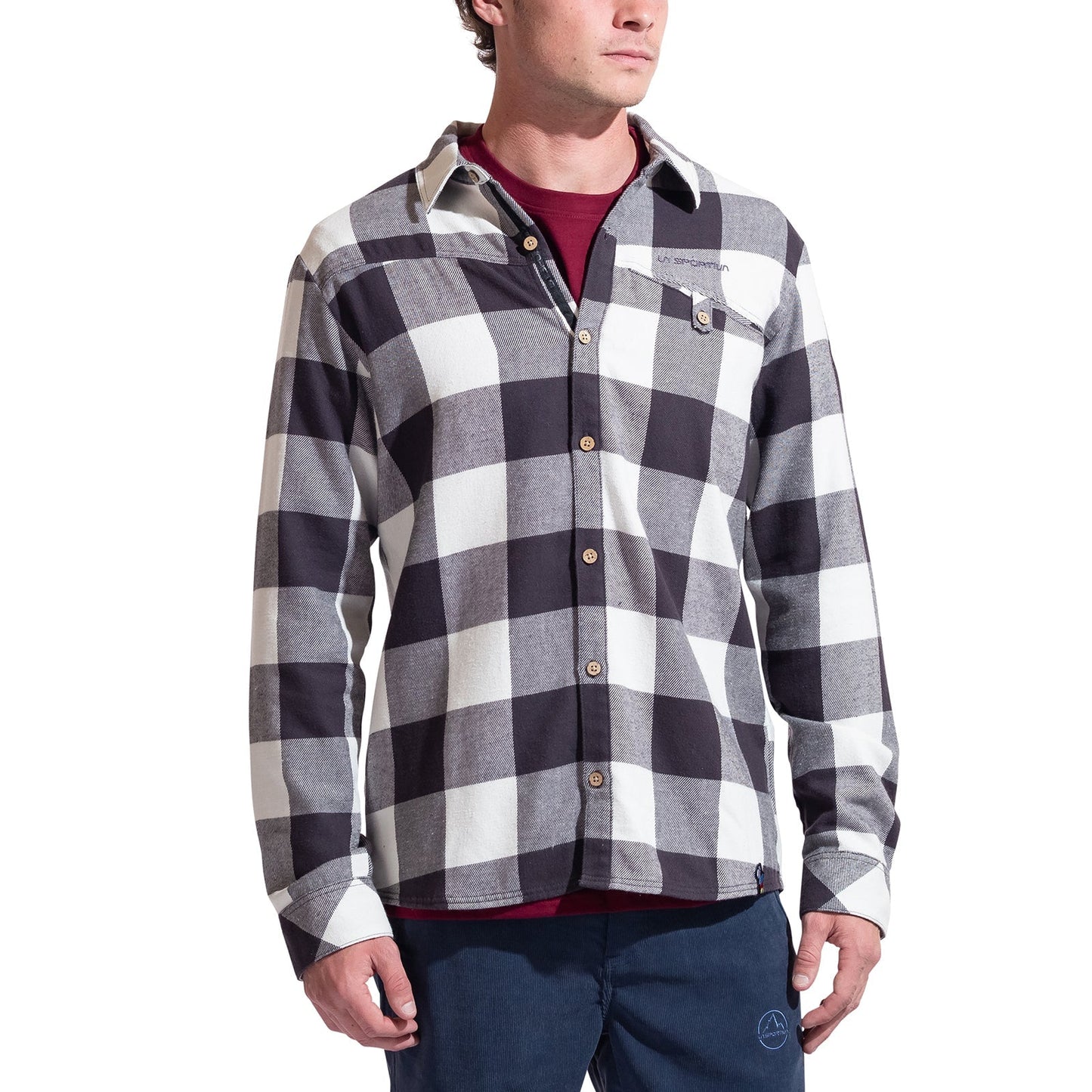 A man wears the La Sportiva Rambler Flannel Shirt in black and white plaid with coconut buttons over a maroon tee and blue pants, standing relaxed against a white background.