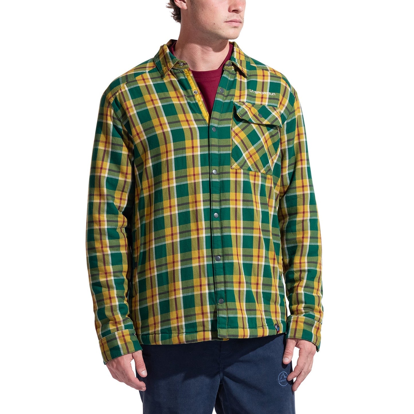 A person wearing the La Sportiva Rambler Lined Flannel Shirt Men's—a green, yellow, and white plaid cotton flannel shirt-jacket with snap buttons—over a red t-shirt and blue pants, standing against a plain white background.