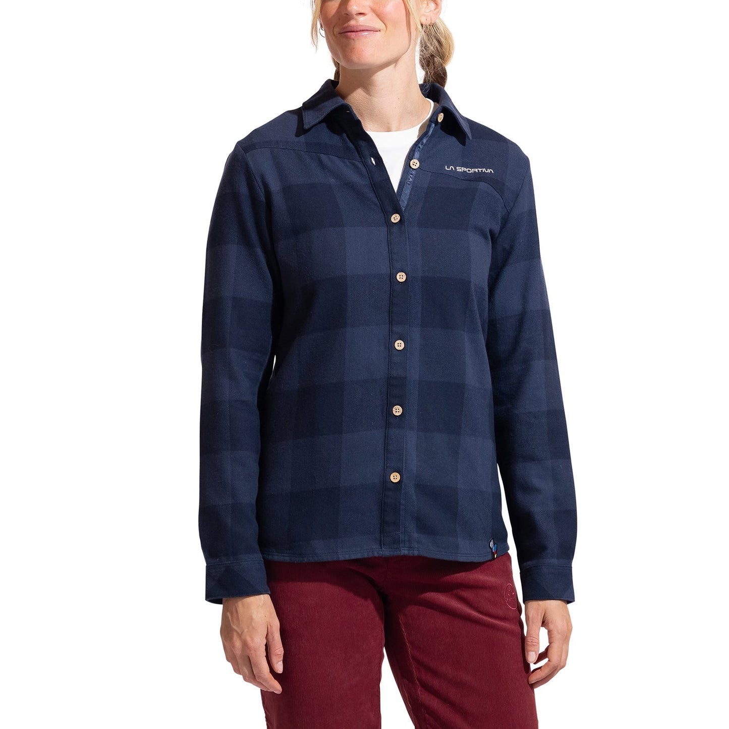 A woman wearing the La Sportiva Rambler Flannel Shirt in dark blue plaid with coconut buttons over a white top and red pants, standing against a white background.