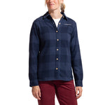 A woman wearing the La Sportiva Rambler Flannel Shirt in dark blue plaid with coconut buttons over a white top and red pants, standing against a white background.