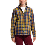 A woman stands and smiles against a white background, wearing La Sportiva's Rambler Lined Flannel Shirt in yellow, navy, and white cotton with snap buttons over a white top and red pants.
