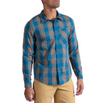 A man wears the La Sportiva Outer Space Long Sleeve Shirt Men's, featuring blue plaid with orange and white stripes and reinforced elbows, along with light tan pants, standing against a white background.