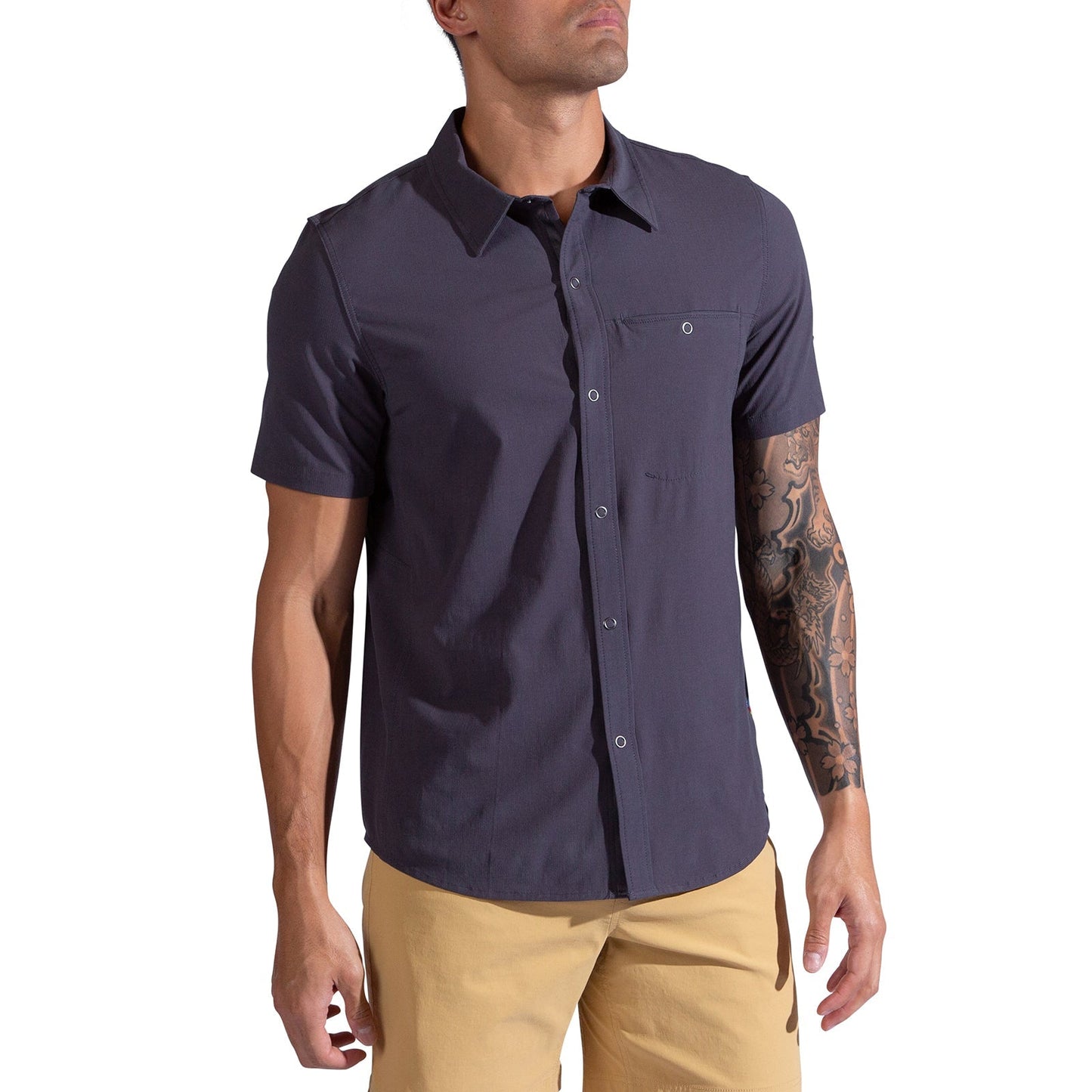 A man wearing the La Sportiva Superfly Short Sleeve Shirt in dark gray and light tan pants stands with his tattooed left arm relaxed, facing slightly to the side against a white background.