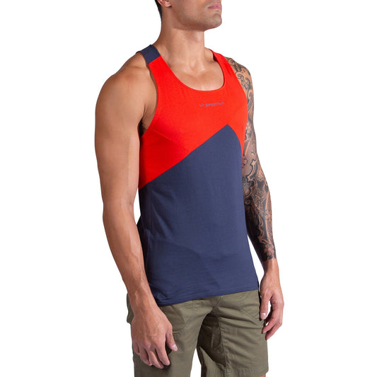 A man models the La Sportiva Dude Tank Men's, a sleeveless top with red and dark blue color blocks, paired with green shorts. A tattoo is visible on his right arm as he stands against a white background.