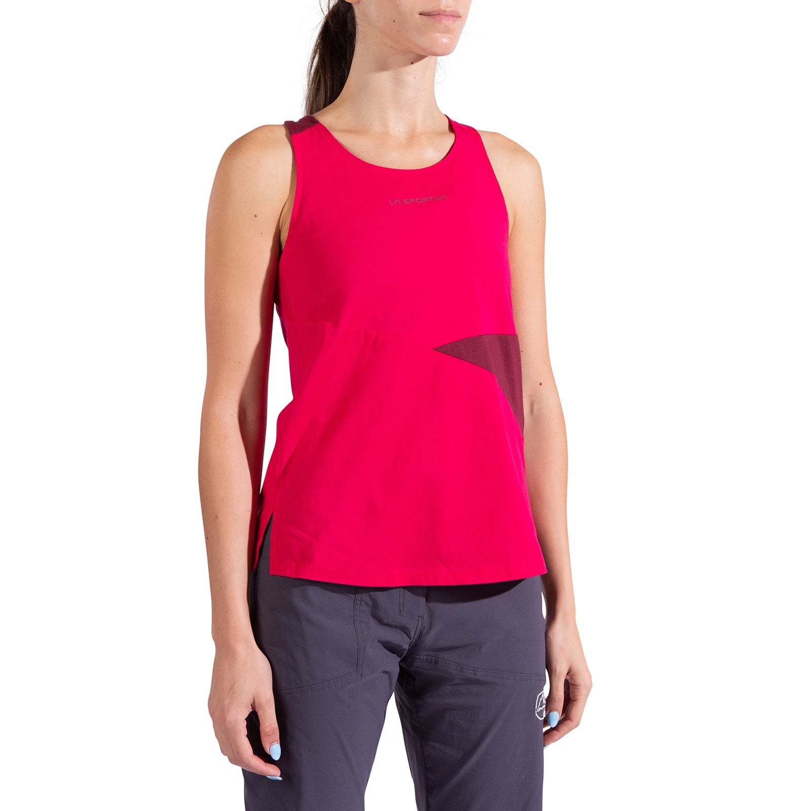 A woman wears the La Sportiva Twist Tank Women's, a sleeveless bright pink organic cotton athletic top, along with dark gray pants. She stands against a white background with her arms relaxed at her sides and head partially out of frame.