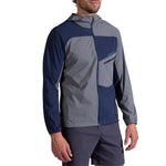 A man wearing the La Sportiva Freeway Wind Jacket Men's, an abrasion-resistant, color-blocked navy blue and gray hooded jacket with a front zipper and chest pocket, paired with dark shorts, stands against a white background.