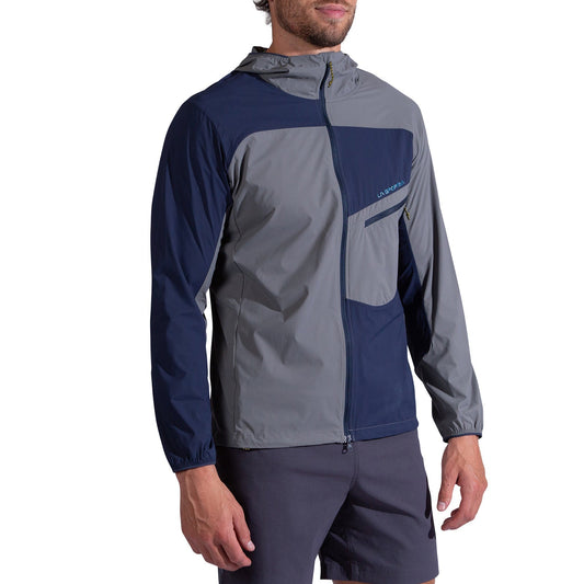 A man wearing the La Sportiva Freeway Wind Jacket Men's, an abrasion-resistant, color-blocked navy blue and gray hooded jacket with a front zipper and chest pocket, paired with dark shorts, stands against a white background.