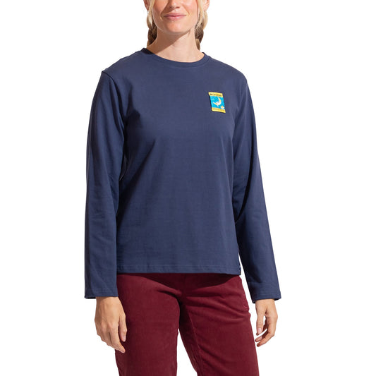 A woman in La Sportiva's Moon Climb Long Sleeve Women's shirt (navy, organic cotton, small colorful chest patch) and burgundy pants stands against a white background.